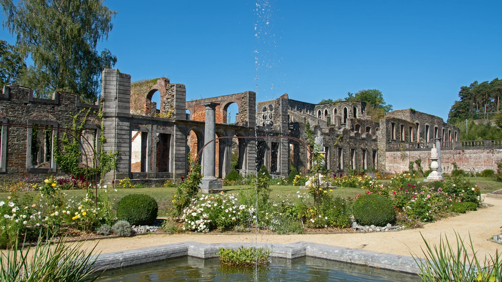 Vue d’ensemble des ruines de l’Abbaye de Villers-la-Ville, avec arches, jardins fleuris et verdure sous un ciel bleu